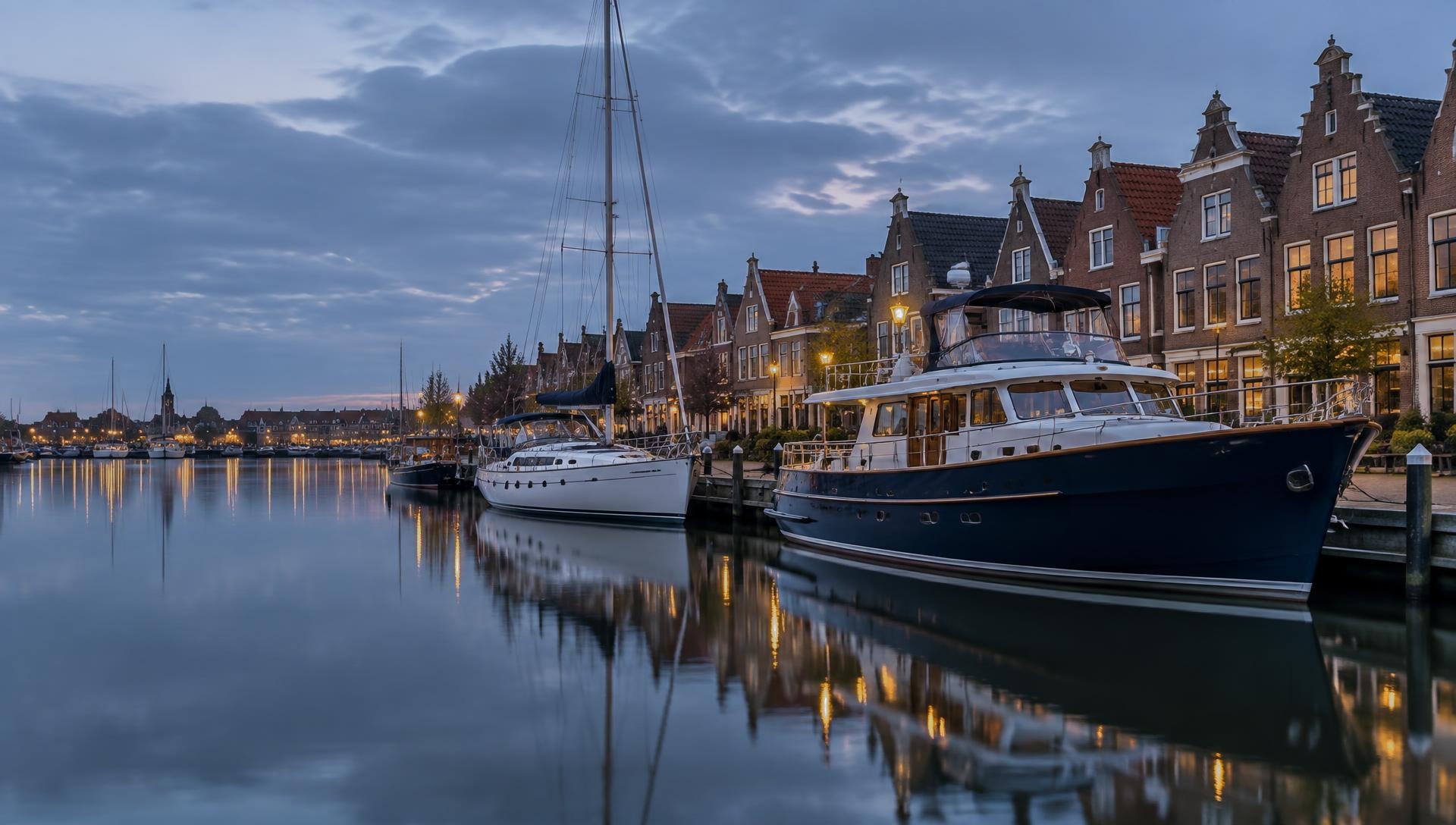 Stahlmotoryacht und Segelyacht in einem niederländischen Hafen vor historischen Giebelhäusern in der Abenddämmerung